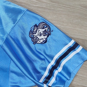 May include: Light blue jersey sleeve with a white and navy stripe design. Features a UNC logo patch with a ram graphic. The jersey has a mesh texture and is made of a shiny, athletic material.