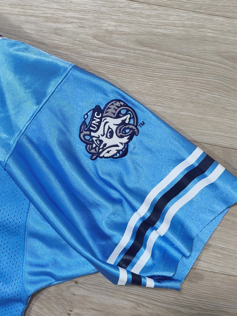 May include: Light blue jersey sleeve with a white and navy stripe design. Features a UNC logo patch with a ram graphic. The jersey has a mesh texture and is made of a shiny, athletic material.