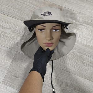 Puede incluir: Sombrero para el sol beige y negro con ala ancha y correa para la barbilla. El sombrero tiene el texto "THE NORTH FACE". Diseñado para la protección solar, es práctico.