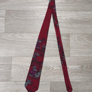 Puede incluir: Una corbata de color rojo intenso con un patrón repetitivo de personajes de ratones de dibujos animados. Los ratones aparecen en varias posturas, con pequeños sombreros azules y ropa a cuadros. La corbata tiene una punta afilada y una forma larga y estrecha, mostrando una base de tela roja vibrante con ilustraciones detalladas y multicolores de los personajes a lo largo de todo el accesorio.