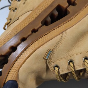 May include: Tan leather boots with a brown rubber sole. The boots feature a lace-up design with metal eyelets and a textured rubber sole. A small blue and white tag is visible on the side.