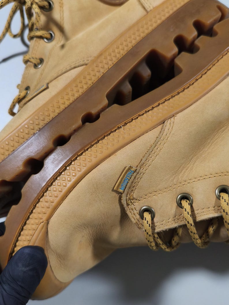 May include: Tan leather boots with a brown rubber sole. The boots feature a lace-up design with metal eyelets and a textured rubber sole. A small blue and white tag is visible on the side.
