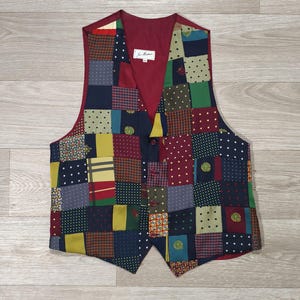 Peut inclure: Un gilet sans manches présentant un motif patchwork composé de divers carrés de tissu dans des tons bleu marine, bordeaux, jaune moutarde et vert. Le vêtement possède une encolure en V, une doublure intérieure bordeaux et une fermeture à boutons.