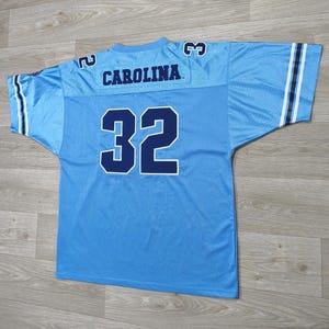 May include: Light blue football jersey with the word "CAROLINA" across the shoulders and the number "32" on the back. The jersey has white and navy blue stripes on the sleeves.