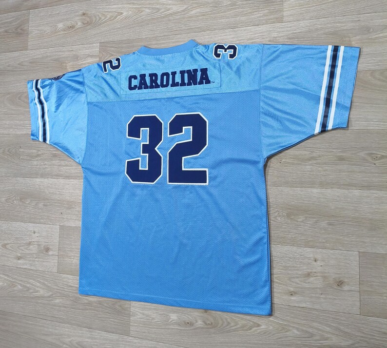 May include: Light blue football jersey with the word "CAROLINA" across the shoulders and the number "32" on the back. The jersey has white and navy blue stripes on the sleeves.