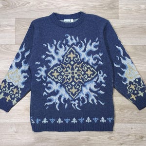 May include: A long-sleeve navy blue crewneck sweater featuring a central diamond-shaped pattern in light blue and cream, surrounded by flame-like motifs. The sleeves and hem also display matching light blue and cream decorative patterns.