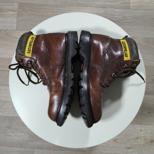 May include: Pair of brown leather work boots with black soles, laces, and the word "CATERPILLAR" printed on a yellow label. The boots are placed on a white surface.