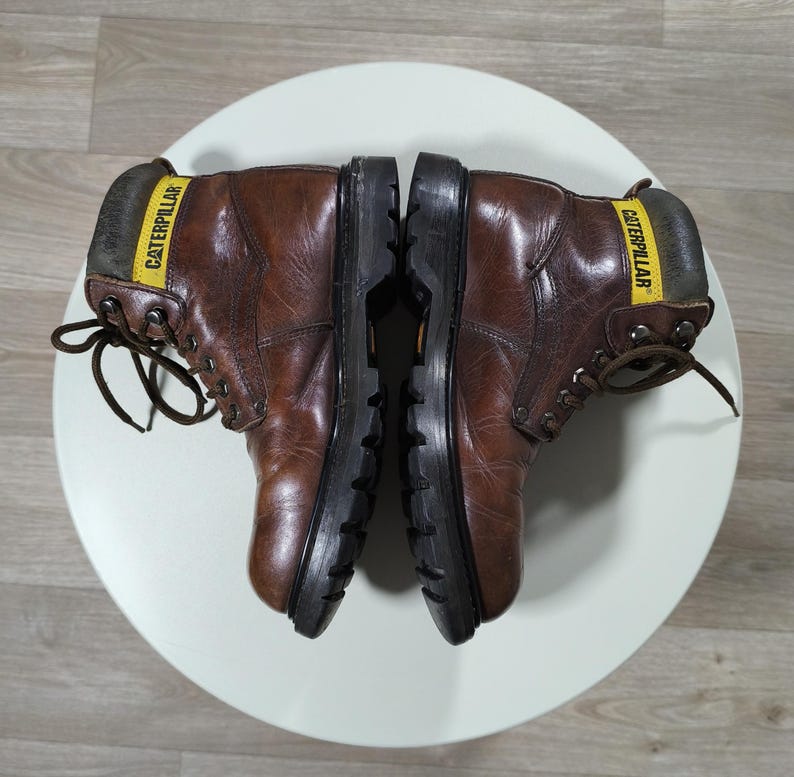 May include: Pair of brown leather work boots with black soles, laces, and the word "CATERPILLAR" printed on a yellow label. The boots are placed on a white surface.