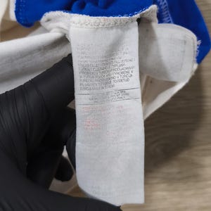 May include: Close-up of a white garment with a blue trim. A white tag with text in multiple languages is attached to the garment. The tag is held by a gloved hand.