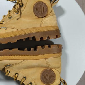 May include: Tan Palladium boots with brown detailing. The boots have a round, embossed logo and a chunky, textured sole. The laces are tan and the boots are made from a robust material, suitable for various terrains.