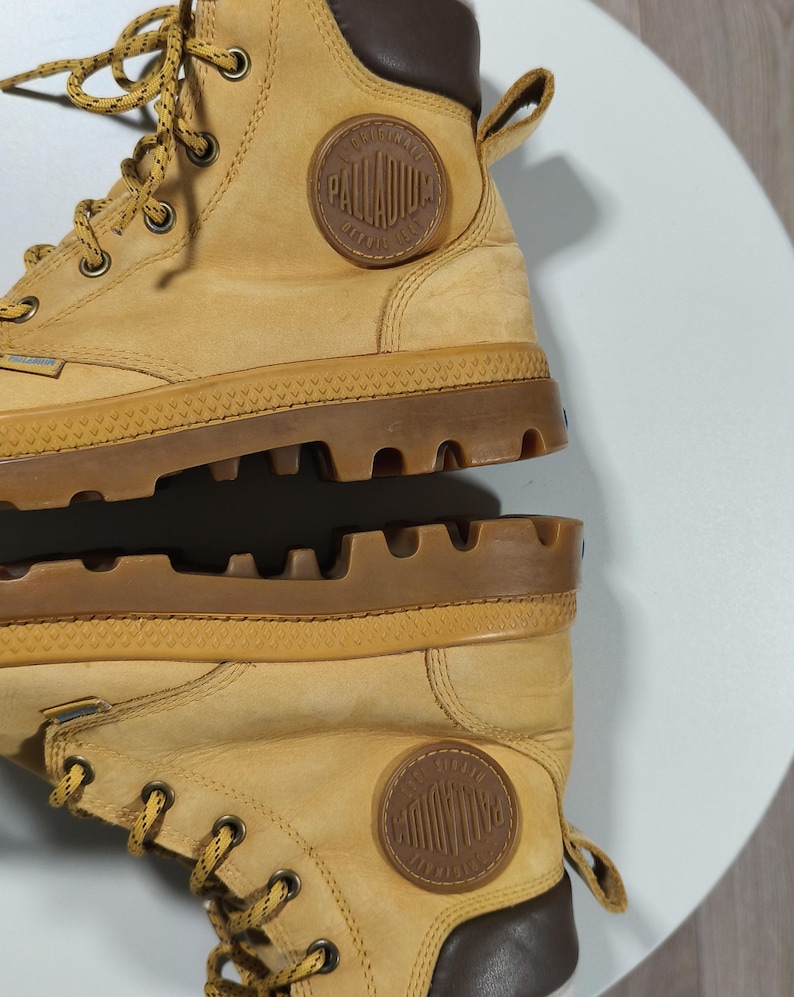 May include: Tan Palladium boots with brown detailing. The boots have a round, embossed logo and a chunky, textured sole. The laces are tan and the boots are made from a robust material, suitable for various terrains.