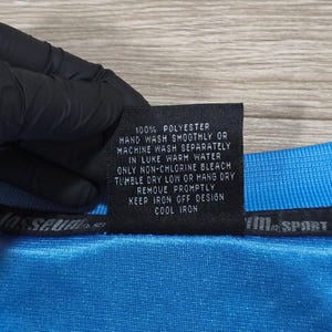 May include: A close-up of a blue polyester garment with a care label. The label states "100% POLYESTER" and provides washing and ironing instructions. The garment has a textured appearance.