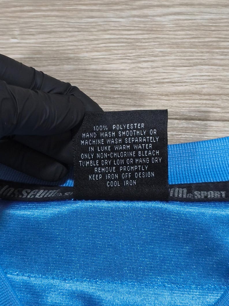 May include: A close-up of a blue polyester garment with a care label. The label states "100% POLYESTER" and provides washing and ironing instructions. The garment has a textured appearance.