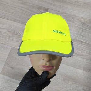 Puede incluir: Gorra de béisbol amarilla neón con visera gris. La palabra "SIEMENS" está impresa en verde en la parte delantera. La gorra se muestra en una cabeza de maniquí.