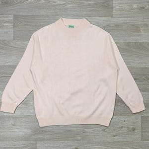 Peut inclure: Un pull rose clair avec un col montant et des manches longues. Le pull a un ourlet et des poignets côtelés. L'étiquette sur le cou indique "Skeans". Le pull est fait d'une matière tricotée douce.