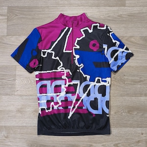 Peut inclure: Maillot de cyclisme à manches courtes avec col zippé. Le maillot présente un motif avec des formes géométriques violettes, bleues et noires, soulignées de blanc. Le maillot est présenté sur une surface en bois.