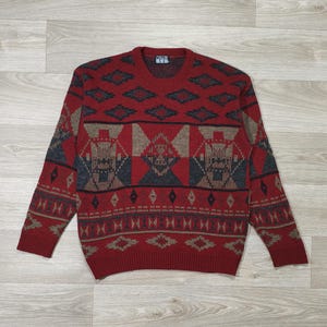 Peut inclure: Un pull rouge avec un motif géométrique en nuances de gris et de beige. Le pull a un col rond et des manches longues. Le motif comprend des losanges et des rectangles, créant un look d'inspiration amérindienne ou du Sud-Ouest.