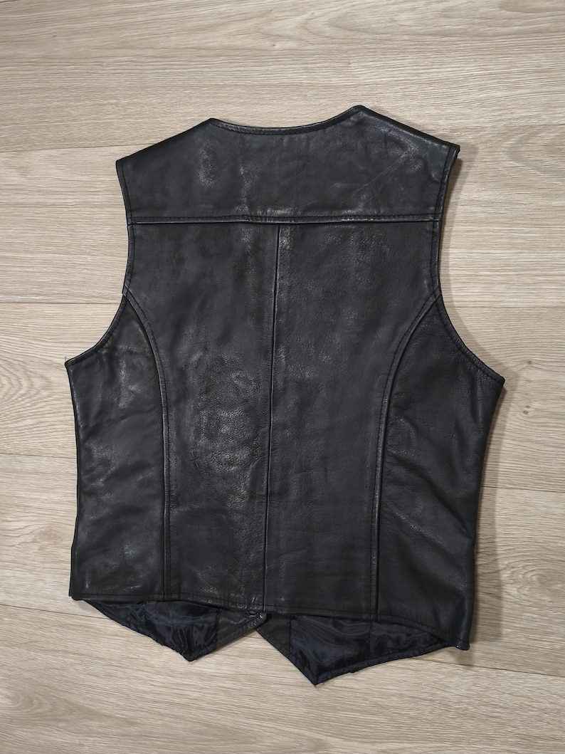May include: Black leather vest with a classic design. The vest features a V-neck, a tailored fit, and a smooth, textured surface. The back of the vest has a panel design with vertical seams. The vest is a versatile piece of apparel.