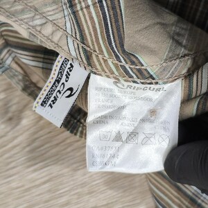May include: A close-up of a tan and brown plaid shirt with a Rip Curl label. The label shows the brand logo and washing instructions. The shirt features a classic plaid pattern with vertical and horizontal stripes.