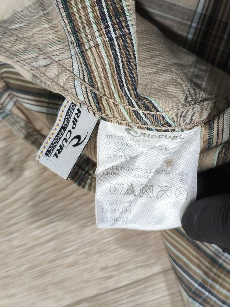 May include: A close-up of a tan and brown plaid shirt with a Rip Curl label. The label shows the brand logo and washing instructions. The shirt features a classic plaid pattern with vertical and horizontal stripes.