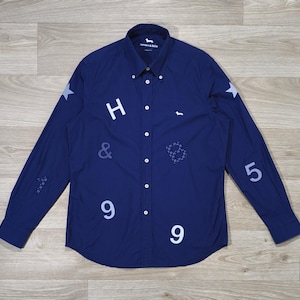 Peut inclure: Chemise bleu marine à manches longues avec des chiffres, des lettres et des étoiles blanches. La chemise a une fermeture boutonnée et un col classique. Le nom de la marque est visible sur le col. Le tissu est lisse.