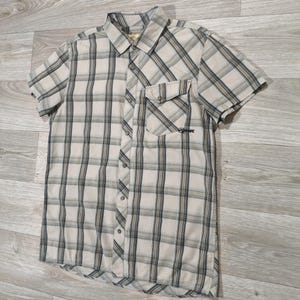 May include: A beige, black, and white plaid short-sleeved shirt. It features a button-down front, a chest pocket, and a collar. The shirt is made of a lightweight material, suitable for casual wear. The shirt has a small logo on the pocket.