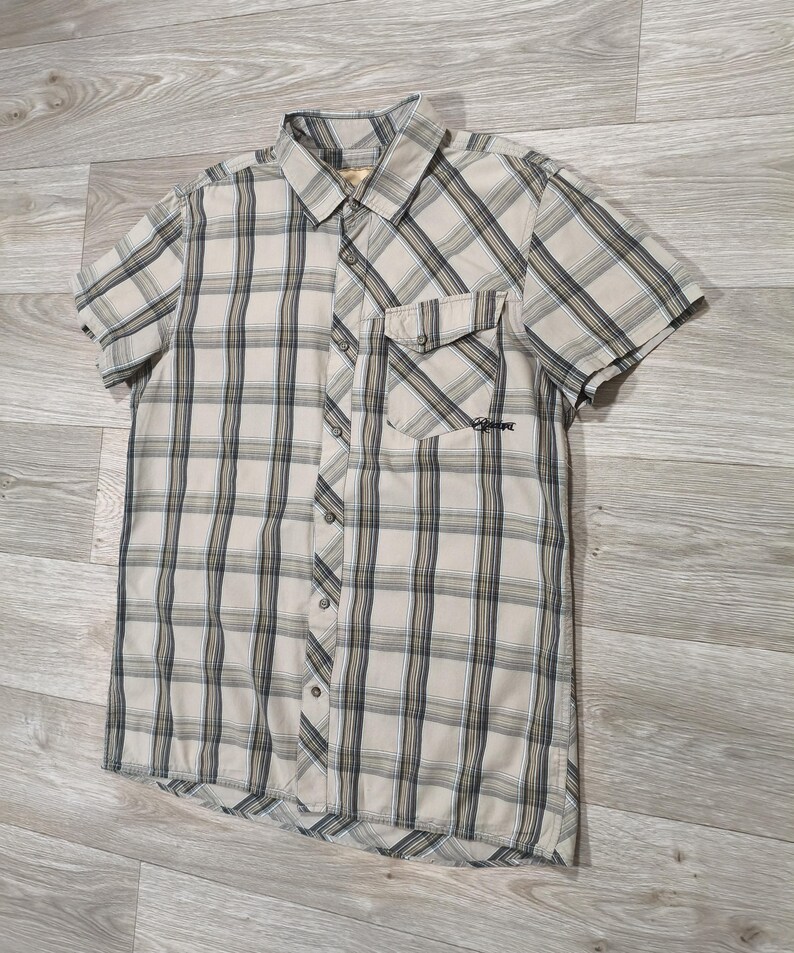May include: A beige, black, and white plaid short-sleeved shirt. It features a button-down front, a chest pocket, and a collar. The shirt is made of a lightweight material, suitable for casual wear. The shirt has a small logo on the pocket.