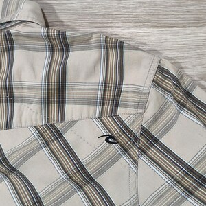 May include: A beige and brown checked shirt. The shirt has a classic plaid design with brown and white stripes. A small black logo is visible on the pocket. The shirt is made of a lightweight material.