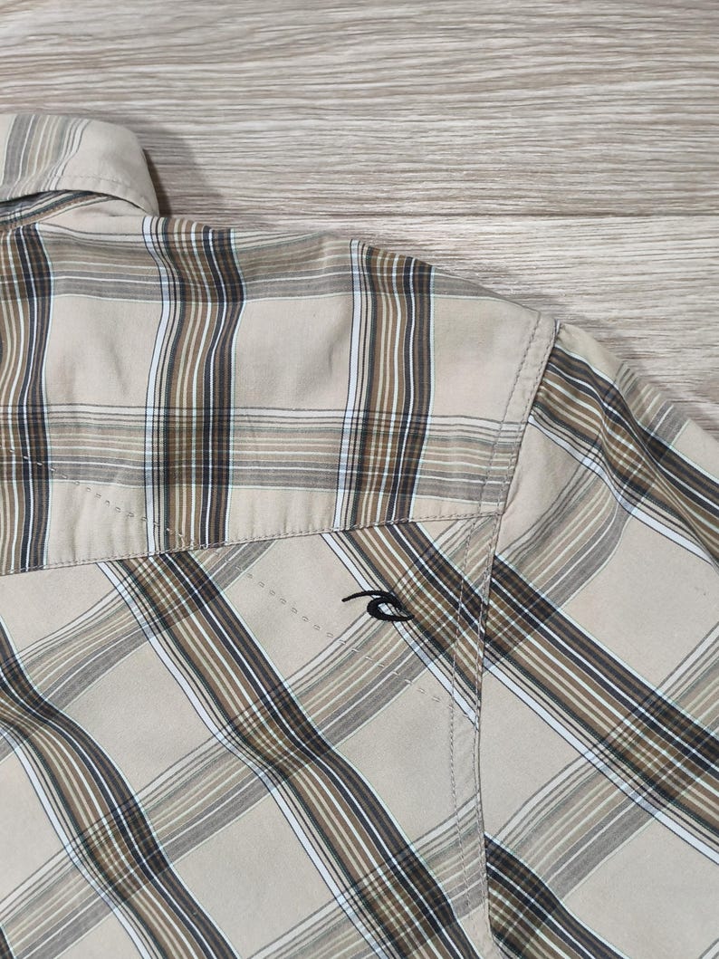 May include: A beige and brown checked shirt. The shirt has a classic plaid design with brown and white stripes. A small black logo is visible on the pocket. The shirt is made of a lightweight material.