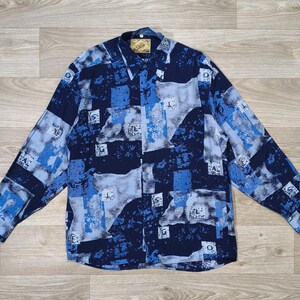 Peut inclure: Une chemise à manches longues avec un motif abstrait bleu, bleu clair et blanc. La chemise a un col et des poignets boutonnés. Le motif comprend diverses formes et motifs de lettres. La chemise est posée à plat sur une surface en bois.