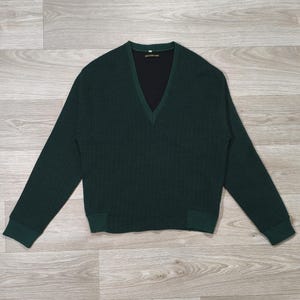 Peut inclure: Un pull col en V vert foncé avec des manches longues et des poignets côtelés. Le pull présente un motif en tricot texturé et une couche intérieure noire visible au niveau de l'encolure. Le vêtement est présenté sur une surface claire imitation bois.