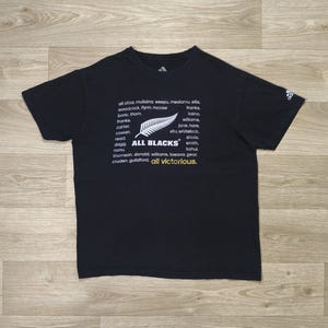 Peut inclure: Un t-shirt noir à manches courtes avec un motif de fougère blanc, le texte ALL BLACKS, une liste de noms et le texte jaune all victorious. Le logo Adidas est imprimé en blanc sur la manche gauche et à l'intérieur du col du t-shirt.