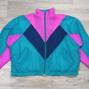 Peut inclure: Veste zippée vintage bleu sarcelle, rose et bleu marine. La veste présente un col montant, deux poches avant et un motif géométrique en blocs de couleurs. C'est un coupe-vent de style rétro.