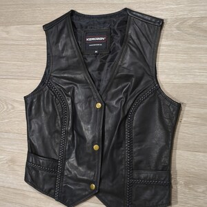 May include: Black leather vest with a V-neck, featuring gold-colored buttons and decorative stitching along the sides. The vest has two front pockets and a label that reads "KOROZAN".