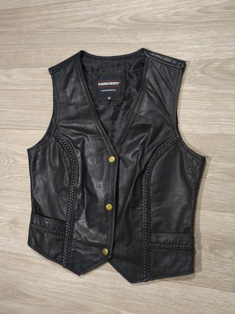 May include: Black leather vest with a V-neck, featuring gold-colored buttons and decorative stitching along the sides. The vest has two front pockets and a label that reads "KOROZAN".