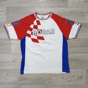 May include: White t-shirt with red sleeves and blue side panels. The shirt features the word "BOBAN" in a bold font, with a checkered pattern. Denim patches are on the sleeves. The shirt has a crew neck and short sleeves.