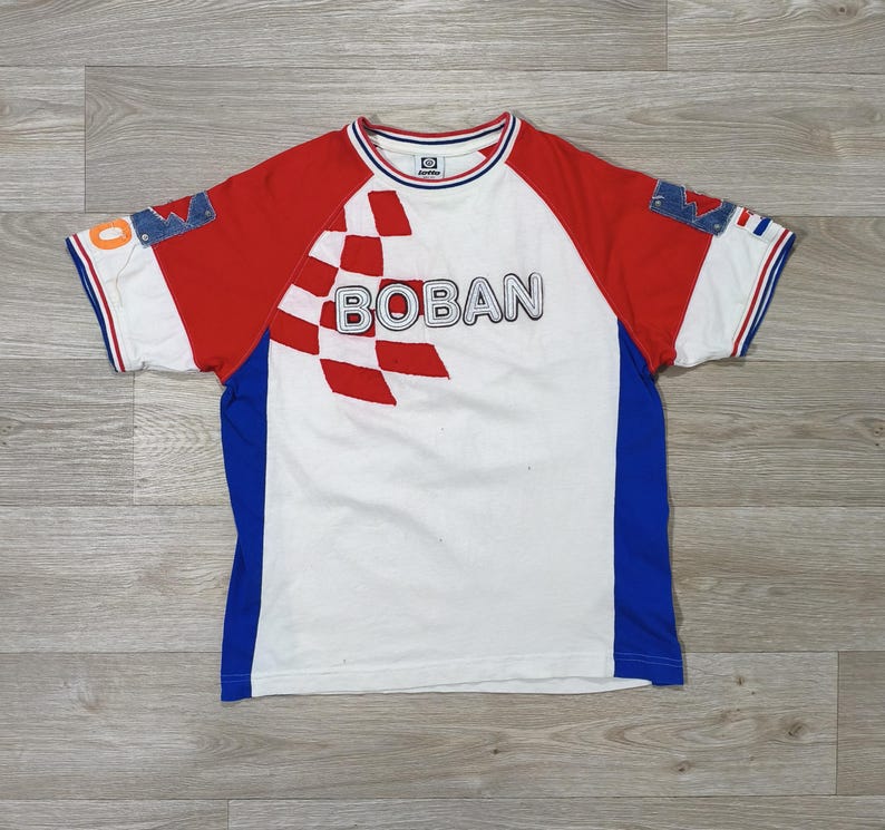 May include: White t-shirt with red sleeves and blue side panels. The shirt features the word "BOBAN" in a bold font, with a checkered pattern. Denim patches are on the sleeves. The shirt has a crew neck and short sleeves.