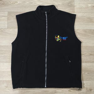 Könnte beinhalten: Schwarze Fleece-Weste mit durchgehendem Reißverschluss und hohem Kragen. Die Weste hat eine kleine gestickte Grafik mit dem Text "Fire 60" in Blau und Gelb. Sie hat zwei Vordertaschen und einen Kordelzug am Saum.