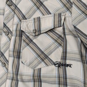 May include: A tan plaid shirt with a chest pocket. The shirt has a classic plaid pattern in shades of tan, brown, and white. The pocket has the embroidered word "Rebel" in black. The shirt has a button-down collar and buttoned cuffs.