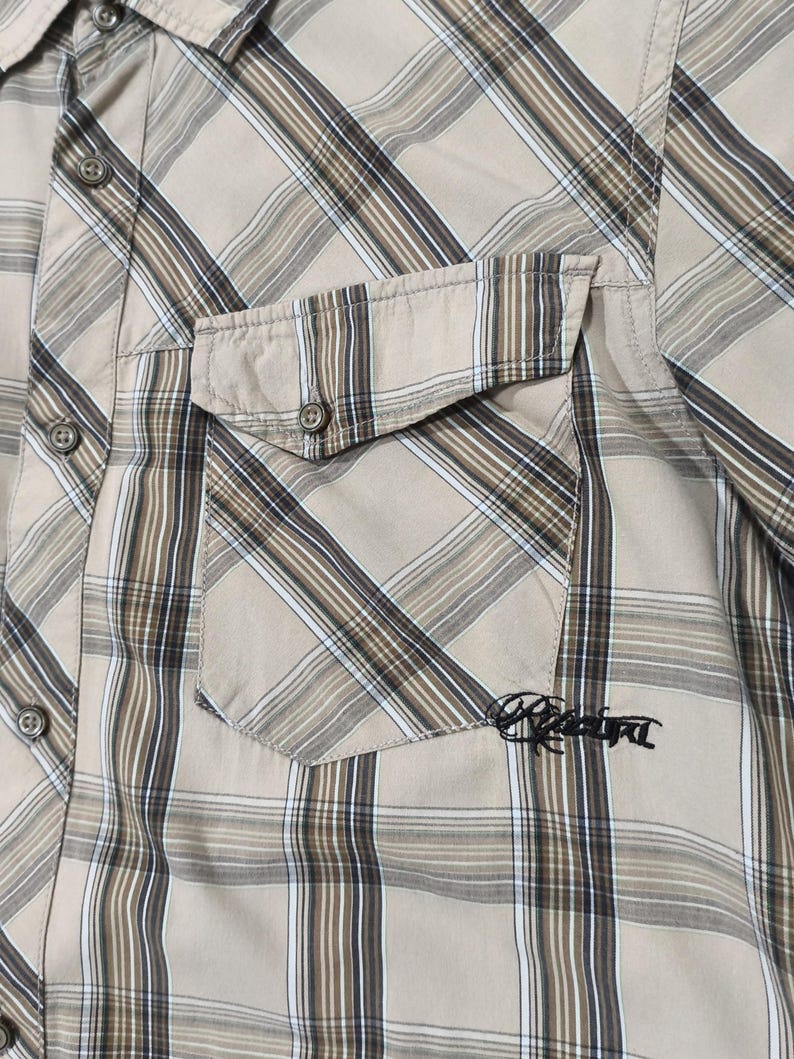 May include: A tan plaid shirt with a chest pocket. The shirt has a classic plaid pattern in shades of tan, brown, and white. The pocket has the embroidered word "Rebel" in black. The shirt has a button-down collar and buttoned cuffs.