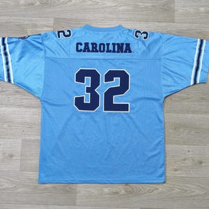 May include: Light blue football jersey. The word "CAROLINA" is across the shoulders, and the number "32" is on the front. The jersey has white and navy blue stripes on the sleeves and the numbers "2" and "3" on the shoulders.