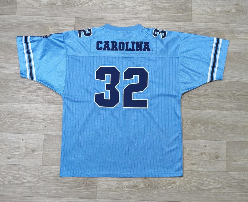 May include: Light blue football jersey. The word "CAROLINA" is across the shoulders, and the number "32" is on the front. The jersey has white and navy blue stripes on the sleeves and the numbers "2" and "3" on the shoulders.