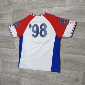 May include: White, red, and blue short-sleeve t-shirt with the number '98' in denim on the back. The shirt has denim patches on the sleeves and a blue stripe down the sides. The collar and sleeves have red and white trim.