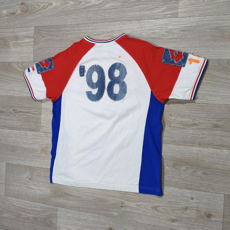May include: White, red, and blue short-sleeve t-shirt with the number '98' in denim on the back. The shirt has denim patches on the sleeves and a blue stripe down the sides. The collar and sleeves have red and white trim.