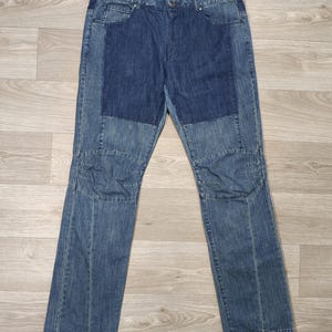 May include: Blue denim jeans with a patchwork design. The jeans have a button closure, pockets, and a straight leg cut. The denim has a medium wash with darker blue patches on the front.
