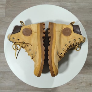 May include: A pair of tan boots with brown details and laces. The boots feature a rubber sole and a logo patch. The boots are displayed on a white surface.