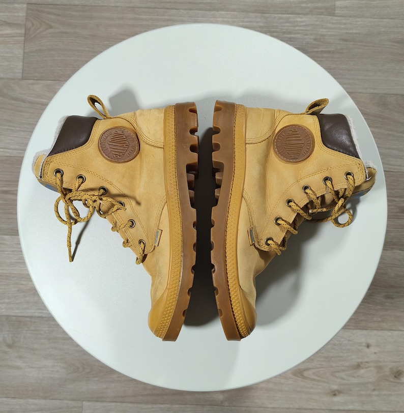May include: A pair of tan boots with brown details and laces. The boots feature a rubber sole and a logo patch. The boots are displayed on a white surface.