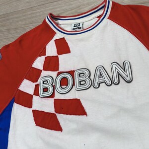 May include: White t-shirt with red sleeves and a red and white checkered design. The word "BOBAN" is across the front in a bold, outlined font. The collar has blue and white stripes, and a small logo is visible.