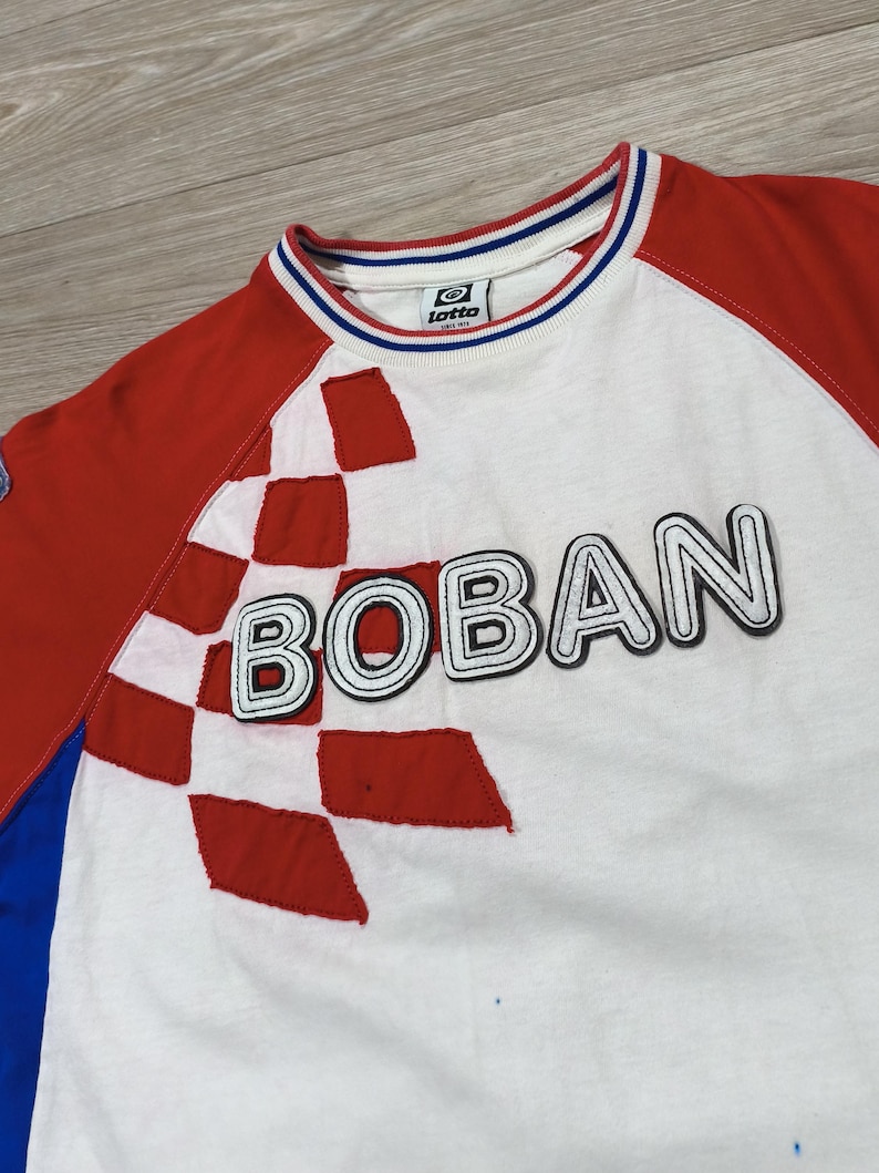 May include: White t-shirt with red sleeves and a red and white checkered design. The word "BOBAN" is across the front in a bold, outlined font. The collar has blue and white stripes, and a small logo is visible.