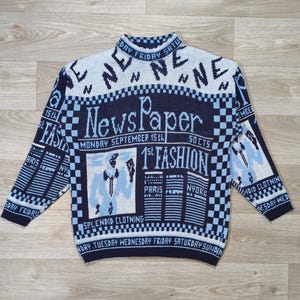 Peut inclure: Un pull bleu marine et bleu clair avec un motif sur le thème du journal. Le pull présente les mots "Newspaper" et "1st Fashion" en grandes lettres, ainsi que d'autres textes et graphiques. Le pull a un col montant et des manches longues.
